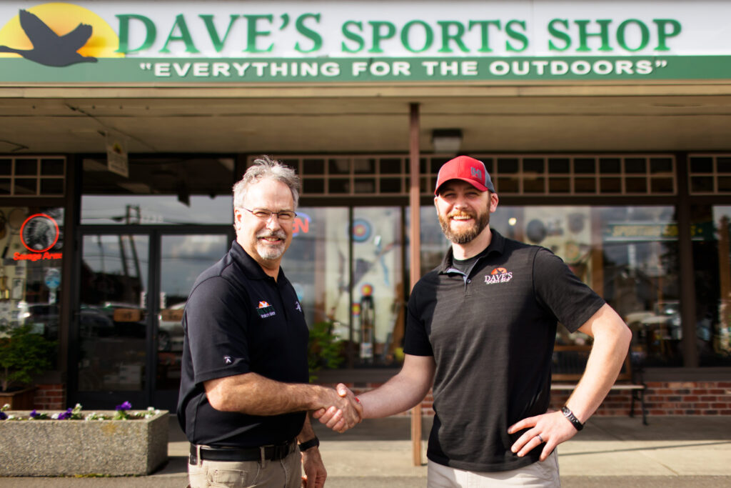 Dave VanderHoek and Will Lathrop shake hands in front of Dave's Sports Shop to commemorate the transition in ownership.