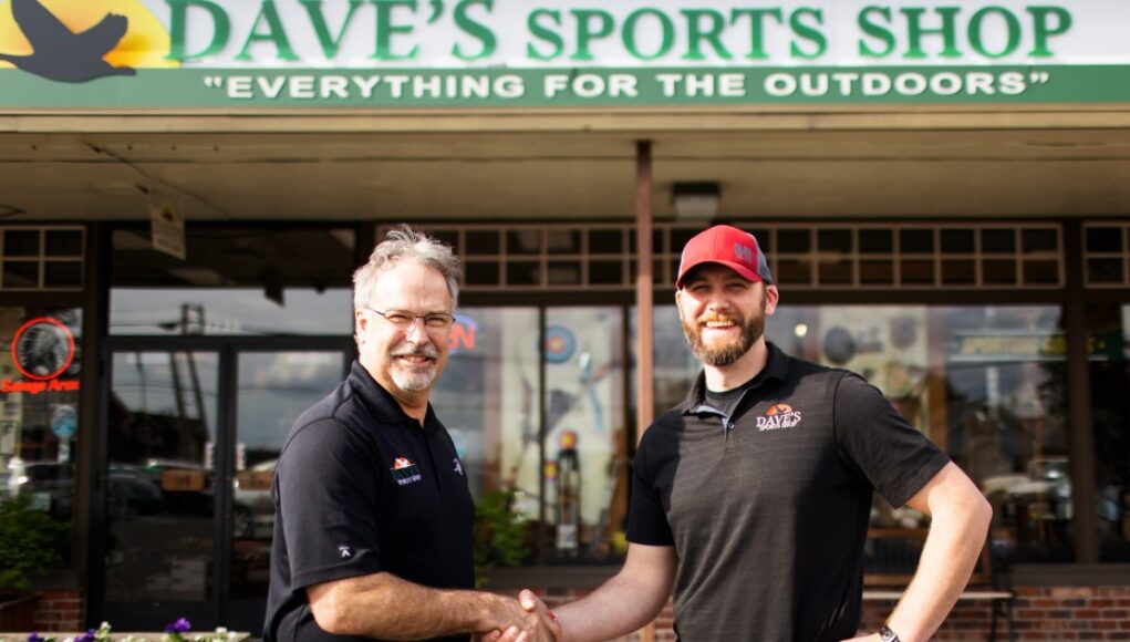 Dave and Will shake hands in front of Dave's Sports Shop to commemorate the sale of the business.